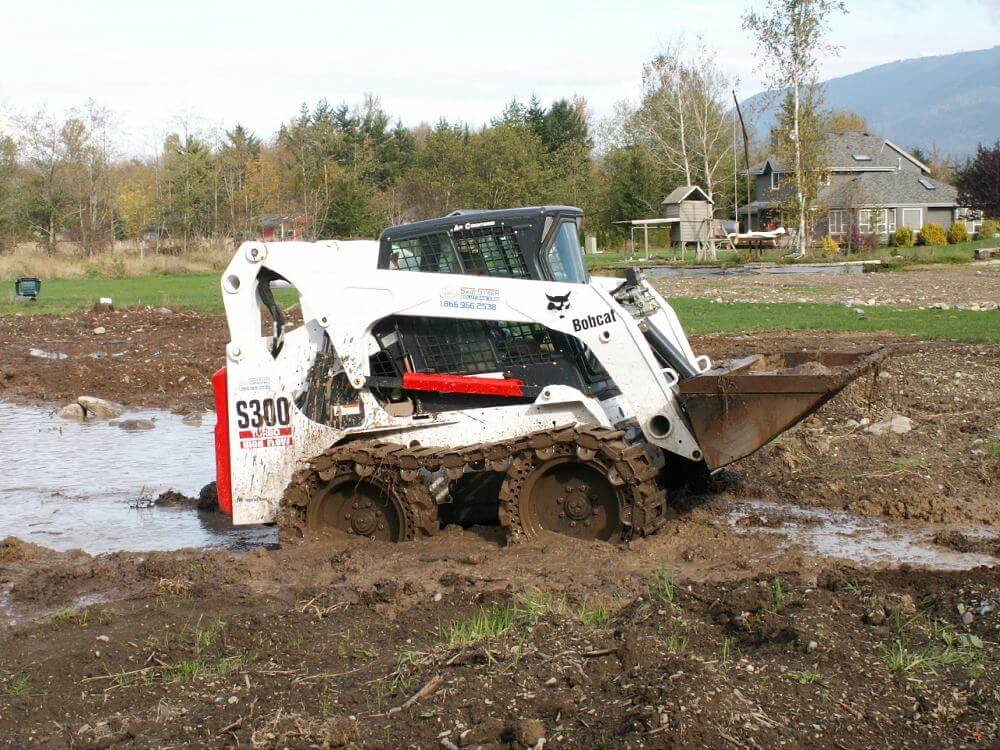A skid steer driving out of a muddy bog with the help of steel tracks attachments